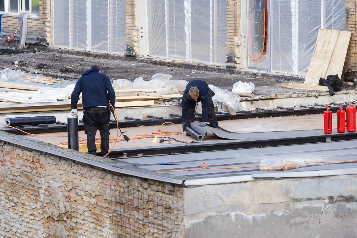 Two construction workers install a black waterproofing membrane and asphalt bitumen shingle on a flat roof