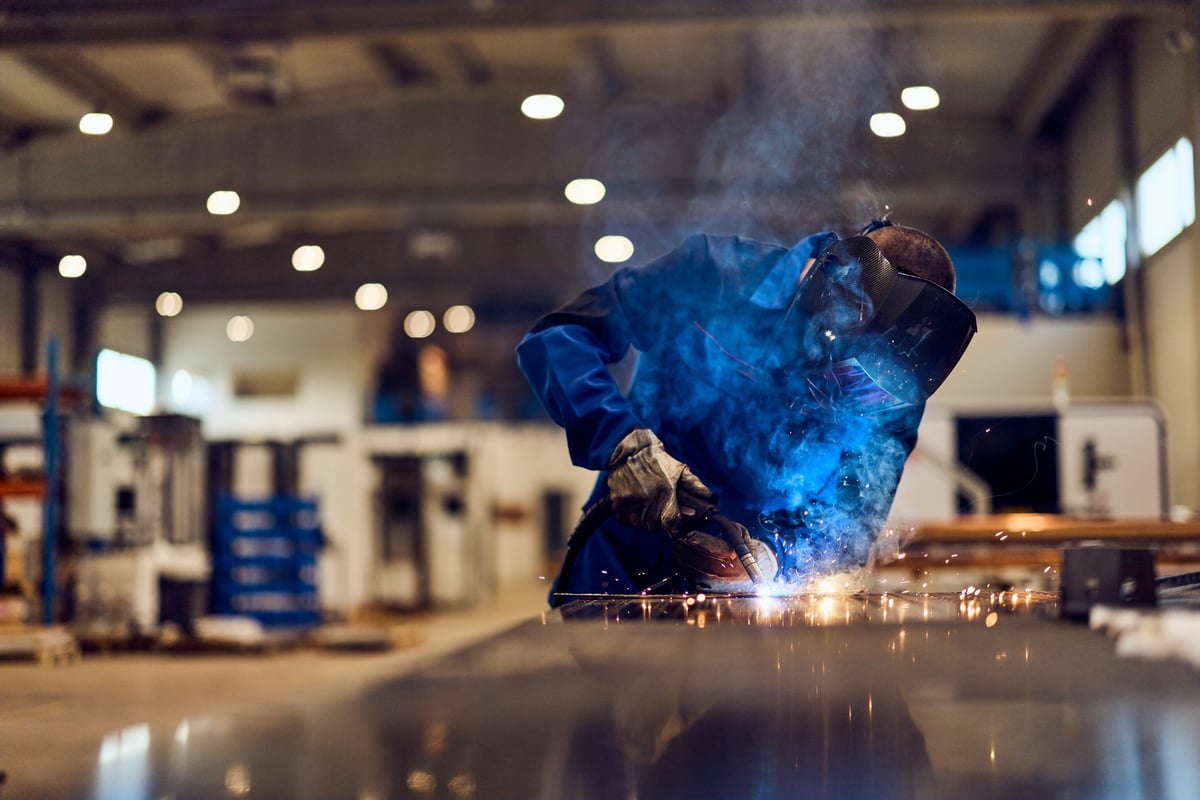 Worker welding metal in a factory workshop, surrounded by sparks and blue smoke.